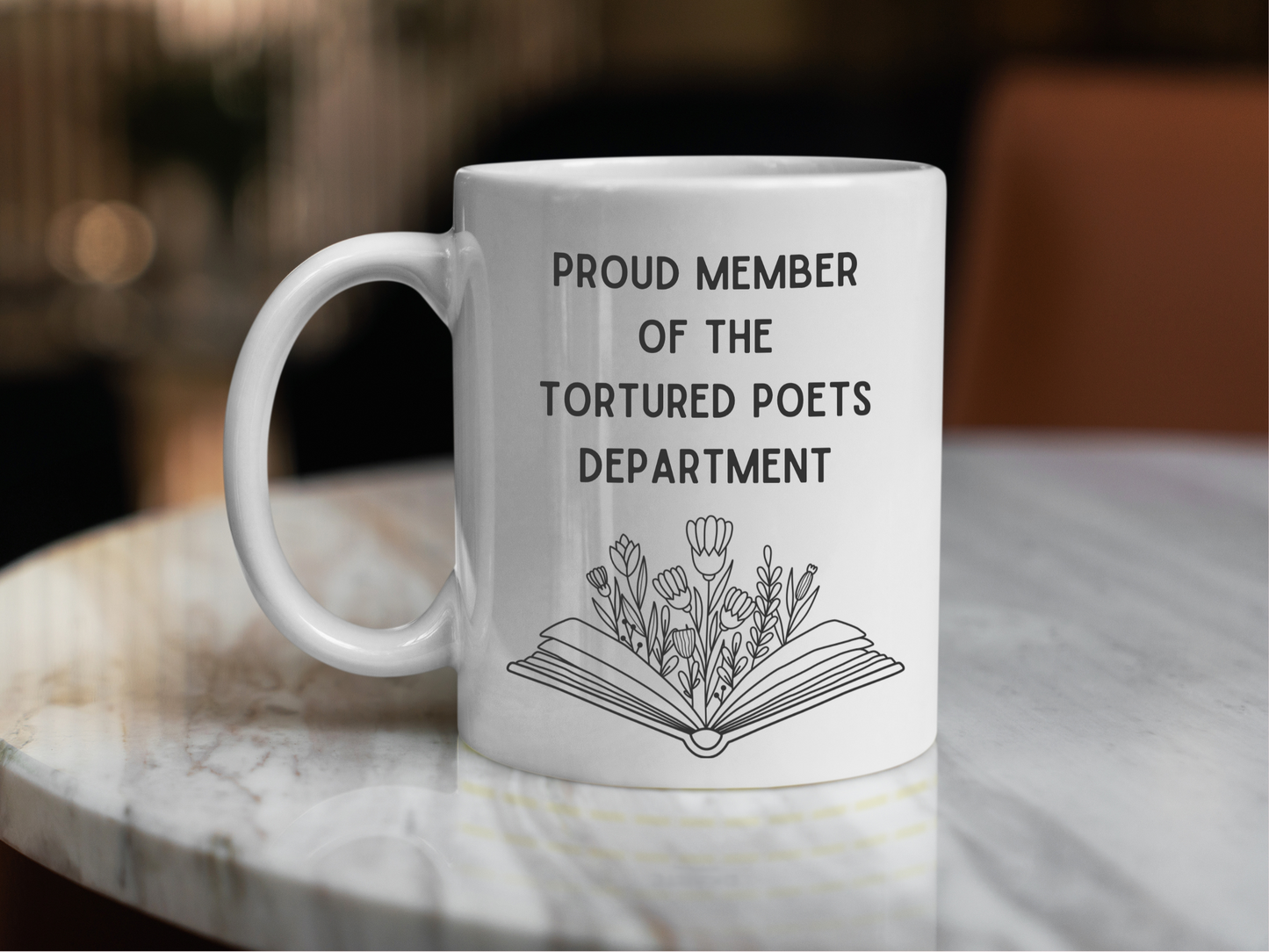 A white ceramic mug with a drawing of a book with flowers, above it is the text Proud member of the Tourtured Poets Department on the front. This is a 10oz mug which is perfect for fans of Taylor Swift. Made and sold by Krissi's Creations.